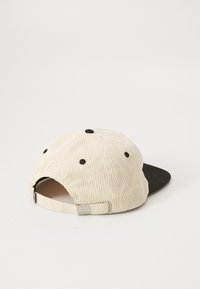 Kordfløyelslue med en beige teksturert krone og en svart flat brem. Har justerbar stropp bak og fem svarte ventilasjonshull.