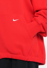 Felpa rossa con cappuccio, tasca anteriore, polsini e orlo a costine. Presenta un piccolo logo bianco Nike sul petto. Il tessuto appare morbido e testurizzato.