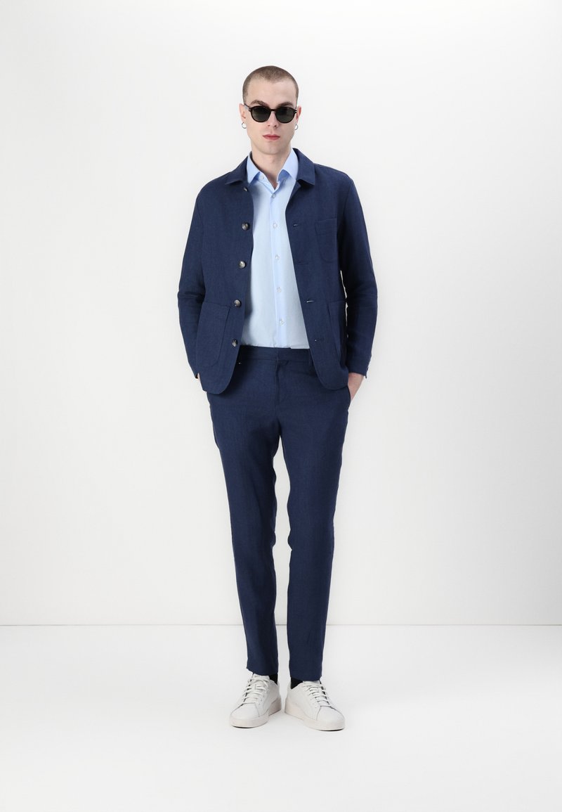 Marineblaues Leinenjackett mit Knöpfen, hellblaues Hemd, marineblaue Hose und weiße Sneakers; Modell trägt schwarze Sonnenbrille und Ohrringe.