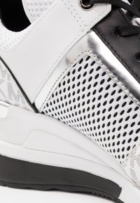 Sneaker featuring black and white synthetic materials, mesh side panels, silver accents, textured surfaces, and a cushioned sole.