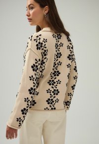 Pull en tricot beige avec broderie florale noire. Il présente des manches longues, un col rond et des bords côtelés. Texture douce et coupe décontractée.