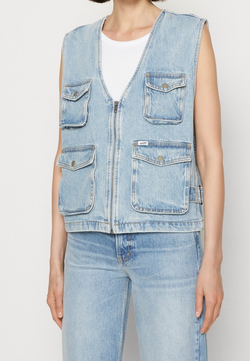 Ärmlös ljusblå denimväst med dragkedja och fyra knappade fickor, bärs över en vit topp med matchande ljusblå denimjeans.