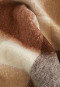 Uldetæppe med en blød, struktureret overflade i en farvepalette af varme brune nuancer, creme og grå i et blandet, abstrakt mønster.