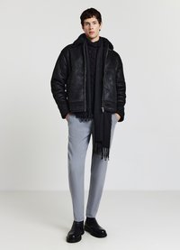 Giacca di pelle nera con collo in shearling, indossata sopra un maglione nero a trecce. Pantaloni grigi sartoriali e una sciarpa nera con frange completano il look.