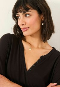 Black, short-sleeved top with a v-neck and five buttons. Gold chain necklace with a round pendant. Model has wavy hair and beige skin.