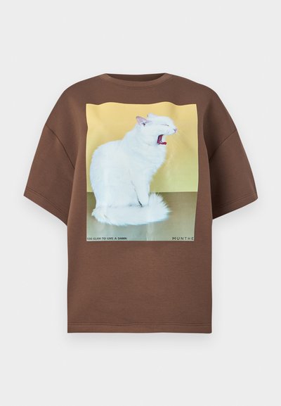 Bruin oversized T-shirt met een grote afbeelding van een gapende witte kat op een gele achtergrond, met tekst onderaan. Zacht katoenen materiaal.