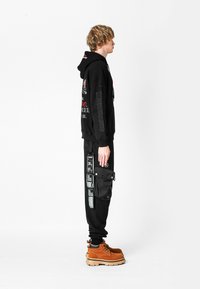 Joven de pie de lado, vistiendo una sudadera gráfica negra, pantalones negros con múltiples parches y un gran bolsillo lateral, y botas de cordones color beige.