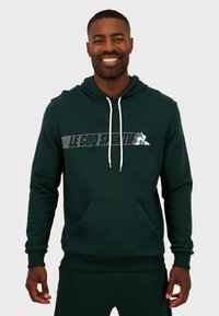 Sudadera verde oscura hecha de material suave, con un bolsillo canguro, capucha con cordón y un logo impreso en blanco en el pecho.