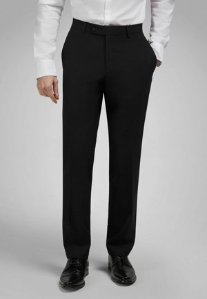 Mann trägt schwarze, maßgeschneiderte Hosen, schwarze Herrenschuhe und ein weißes, langärmeliges Hemd mit Knöpfen, die Hände in den Taschen, steht vor grauem Hintergrund.