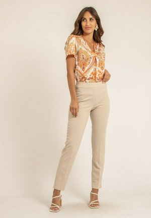 Blouse à manches courtes en crème avec un motif paisley orange et vert, associée à un pantalon slim beige et des sandales à talons blanches.