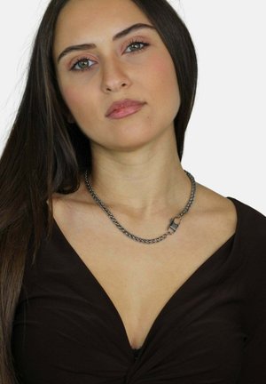 Collana in argento con segmenti interconnessi e un dettaglio di chiusura, indossata su un top marrone scuro. Texture liscia, design minimalista.