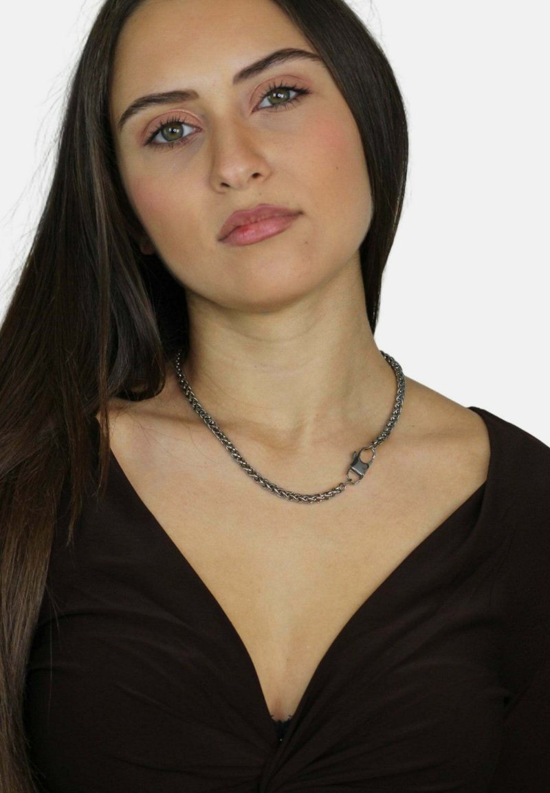 Collana in argento con segmenti interconnessi e un dettaglio di chiusura, indossata su un top marrone scuro. Texture liscia, design minimalista.