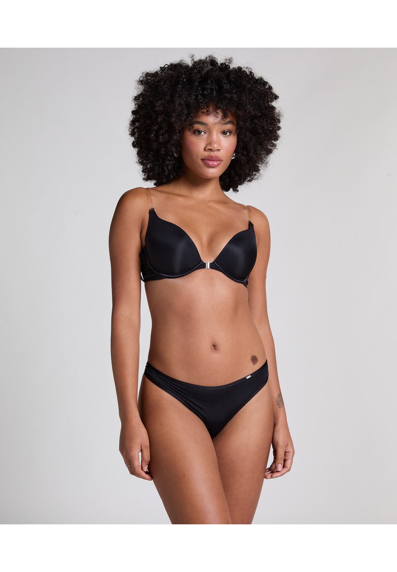 Woman with curly hair wearing a black bra and matching low-rise briefs against a plain gray background.