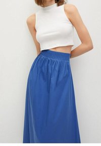 Donna che indossa un top corto senza maniche bianco e una gonna blu a vita alta e plissettata contro uno sfondo chiaro e uniforme.