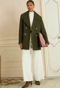 Dvogubna maslinasto zelena kaput s velikim ovratnikom, bijelim gumbima i srebrnim detaljima, uparen s bijelim topom i širokim hlačama.