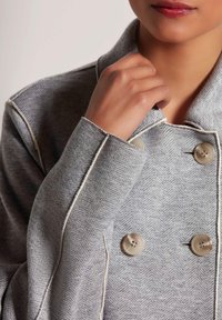 Grijze visgraatjas met een dubbelgeknoopte ontwerp, contrasterende witte stiksels en grote knopen aan de voorkant. Zacht, gestructureerd stof.