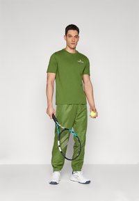 Maslinastozeleni kratki rukav T-shirt s logotipom, u paru s odgovarajućim zelenim trenirkama i bijelim sportskim tenisicama, drži tenisku reketa i lopticu.