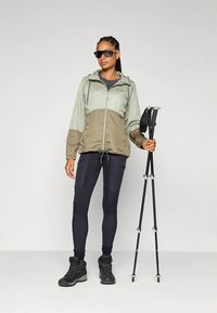 Lichtgewicht, tweekleurig jack in mintgroen en kaki, met een ritssluiting aan de voorkant en een capuchon. Gecombineerd met zwarte leggings en stevige zwarte wandelboots.