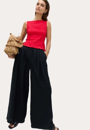 Kobieta w czerwonym, bezrękawnym, teksturowanym topie oraz czarnych, szerokich spodniach, trzymająca warstwową słomianą torebkę z frędzlami.