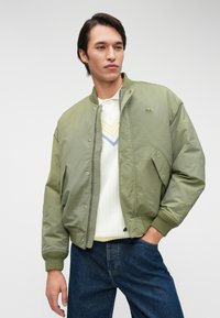 Λαδί bomber jacket με κλείσιμο φερμουάρ και κουμπιά, ριμπ μανσέτες και μια μικρή λογότυπη πινακίδα στο αριστερό στήθος. Φορεμένο πάνω από ένα λευκό πλεκτό τοπ.