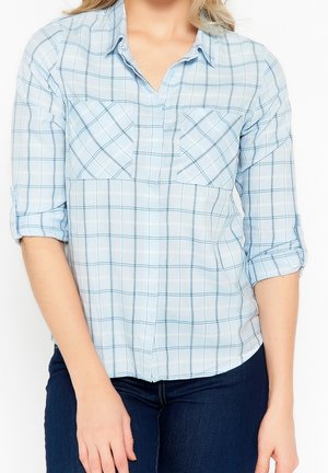 Kvinde iført en lyseblå ternet knap-op skjorte med smalle ærmer og mørkeblå jeans, stående med armene afslappet ned langs siderne.