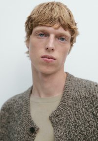 Brauner strukturierter Cardigan mit schwarzen Knöpfen, getragen über einem hellbeigen Hemd. Das Modell hat kurze gewellte blonde Haare und einen neutralen Gesichtsausdruck.