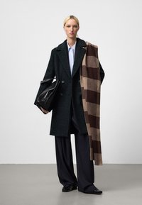 Cappotto doppiopetto verde scuro in tessuto testurizzato, abbinato a una sciarpa a quadri marrone. Il modello tiene una borsa in pelle nera.