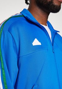 Giacca a zip blu con logo Adidas bianco, dotata di strisce verdi e rosse sulle spalle e un tessuto liscio e strutturato.
