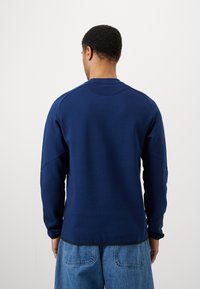 Temno modra majica s dolgimi rokavi, z ribanim izrezom in gladko teksturo. Ima okroglo obliko hrbtnega roba in subtilne detajle šivov.