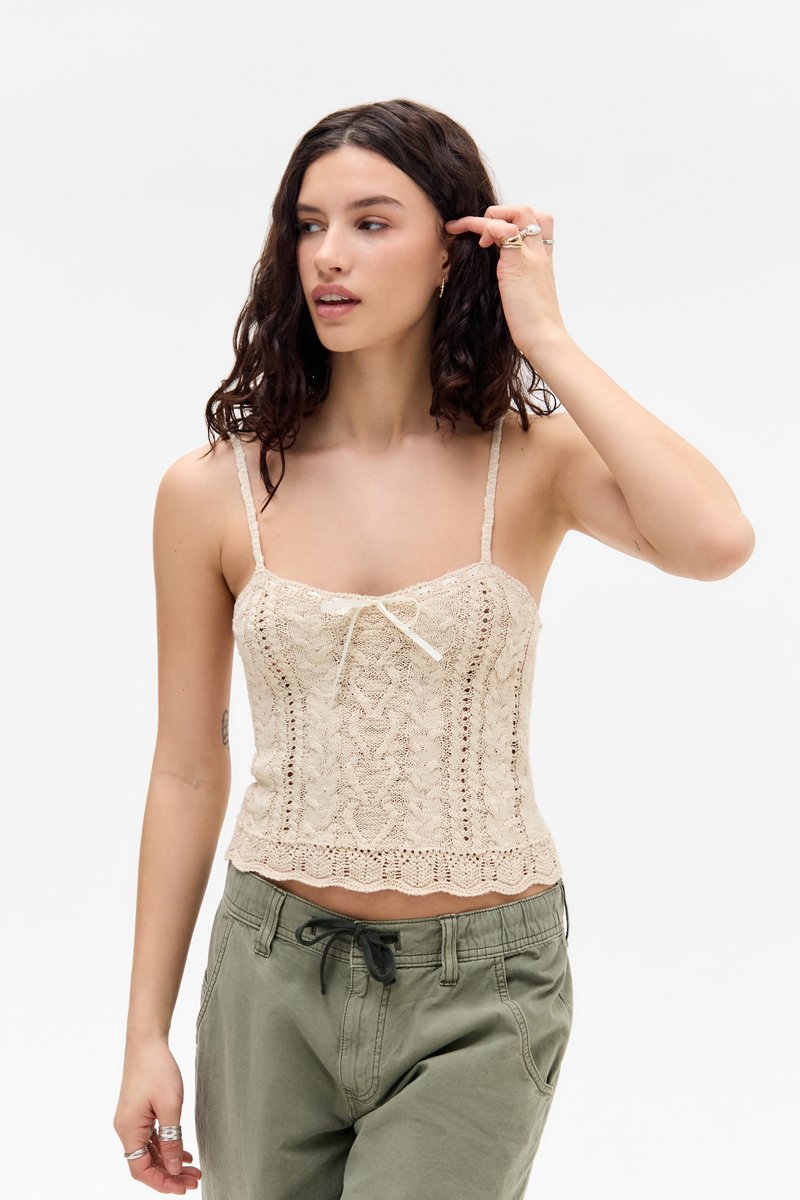 Bež pleteni crop top z nastavljivimi naramnicami, ki ima na sprednji strani detajl s pentljo in valovit rob. Tkanina je teksturirana.
