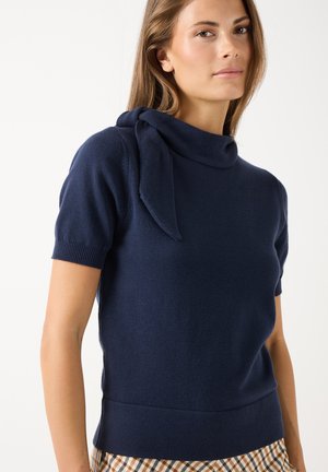 Frau trägt einen dunkelblauen Kurzarmpullover mit einem Stoffbindedetail am Ausschnitt und karierte Hosen vor einfachem Hintergrund.