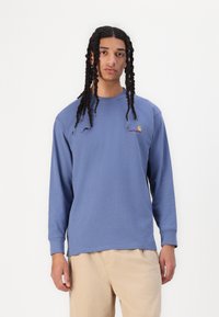 T-shirt en coton bleu à manches longues avec un col rond, arborant un petit logo jaune et rouge sur la poitrine gauche, porté avec un pantalon beige.