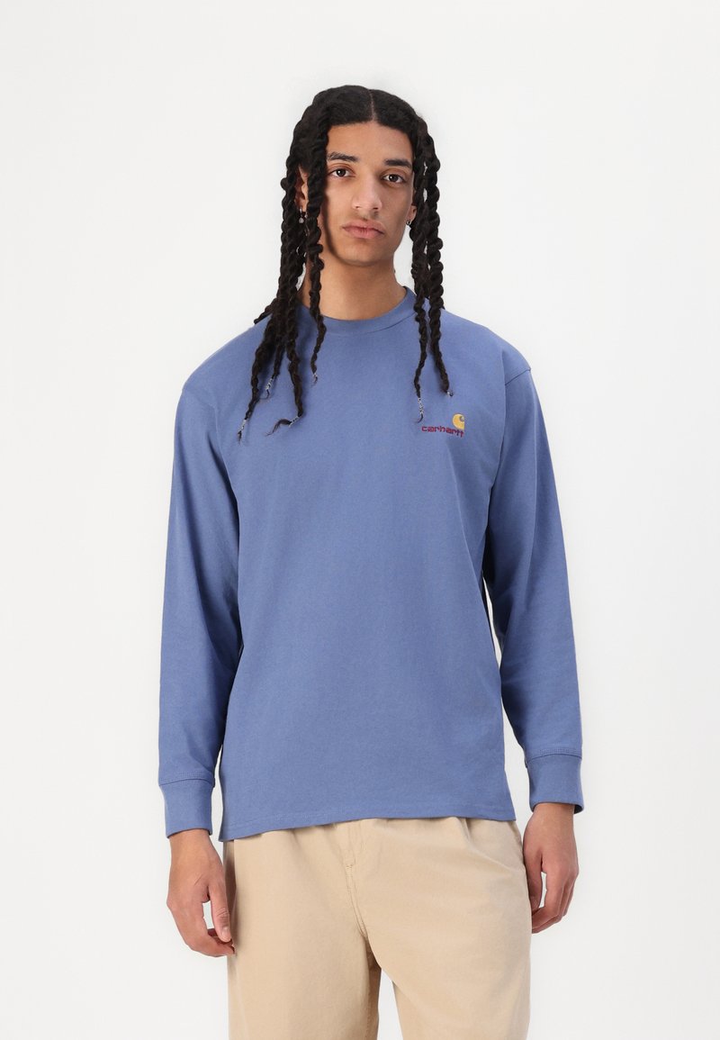 T-shirt en coton bleu à manches longues avec un col rond, arborant un petit logo jaune et rouge sur la poitrine gauche, porté avec un pantalon beige.