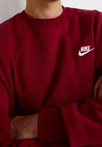 Person klädd i en vinröd Nike-tröja med armarna i kors, som visar den vita Nike-logotypen på bröstet.