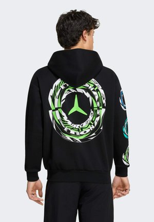 Mężczyzna w czarnej bluzie z kapturem z dużym zielono-białym okrągłym logo "Mercedes-Electronic" na plecach oraz mniejszymi logo na rękawach.