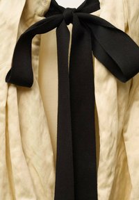 Cream fabric with vertical pleats features a central black fabric tie. The tie is flat, textured, and long, creating a contrast with the smooth background.