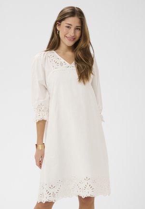 Mladá žena s dlhými vlasmi, oblečená v bielych šatách po kolená s čipkovými detailmi na rukávoch, výstrihu a leme, usmieva sa na jednoduchom pozadí.