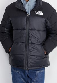 Persona vestida con una chaqueta acolchada negra con el logo de The North Face, sudadera azul clara y jeans azules, de pie con las manos en los bolsillos de la chaqueta.
