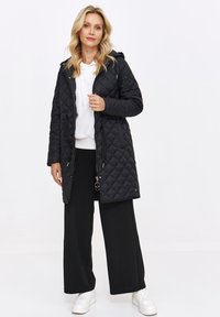 Pikowana czarna kurtka z kapturem, z zatrzaskami i sznurkiem w pasie, połączona z białym hoodie i czarnymi spodniami o szerokich nogawkach.