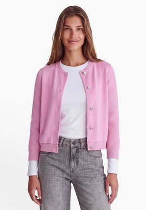 Jonge vrouw met lang bruin haar die een roze vest met knopen draagt over een wit shirt en grijze spijkerbroek, staand tegen een witte achtergrond.