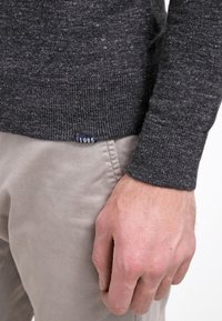 Main à côté d'un pantalon beige clair avec un pull en maille gris foncé montrant une petite étiquette "1985" près de l'ourlet.