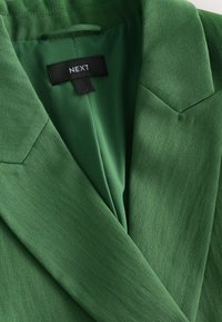 Blazer vert sur mesure avec revers crantés et étiquette de marque noire "NEXT" cousue à l'intérieur du col.