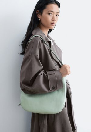 Jeune femme portant un manteau marron oversize, tenant un sac à épaule vert clair sur un fond uni clair.