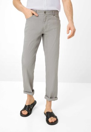 Mann trägt hellgraue hochgekrempelte Hose und schwarze Sandalen mit Kreuzriemen, steht mit einer Hand in der Tasche.