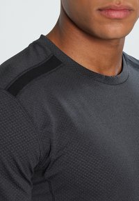 Zbliżenie na osobę w ciemnoszarej, wzorzystej koszulce sportowej z czarnymi detalami na ramionach, pokazujące szyję i częściowo twarz.