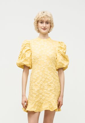 Ung person med kort krøllet blondt hår iført en gul tekstureret kjole med oppustede ærmer og blomsterprint, stående mod en ensfarvet baggrund.
