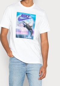 Vit t-shirt med kort ärm som har en färgglad grafik av en späckhuggare, texten "NIKE AIR" och Nike-logotypen. Avslappnat snitt med rund halsringning.