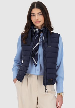 Frau trägt navyblaue gesteppte Weste über hellblauer Bluse, gemusterten Halstuch, beigefarbenen Hosen und goldener Armbanduhr, steht vor einfarbigem Hintergrund.