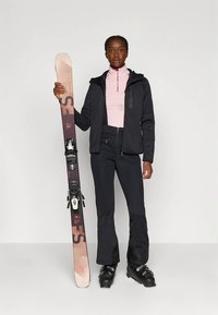 Pessoa vestida com um fato de ski, segurando um único ski em pé, usando botas pretas, calças, uma jaqueta e uma parte superior rosa clara com fecho contra um fundo liso.