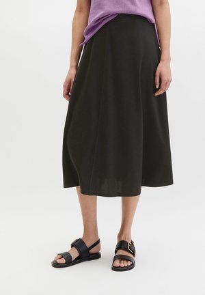 Kobieta ubrana w czarną spódnicę midi, lawendową bluzkę i czarne sandały na otwartym palcu z złotą klamrą, stojąca na białym tle.